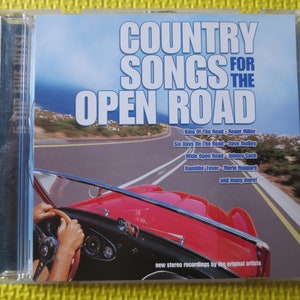 Op de afbeelding: Een rode cabriolet rijdt over een kronkelende weg met een blauwe lucht en de oceaan op de achtergrond. De CD-hoes heeft de tekst "Country Songs for the Open Road" met een lijst van nummers en artiesten, waaronder "King of the Road - Roger Miller", "Six Days on the Road - Dave Dudley", "Wide Open Road - Johnny Cash" en "Ramblin' Fever - Merle Haggard".