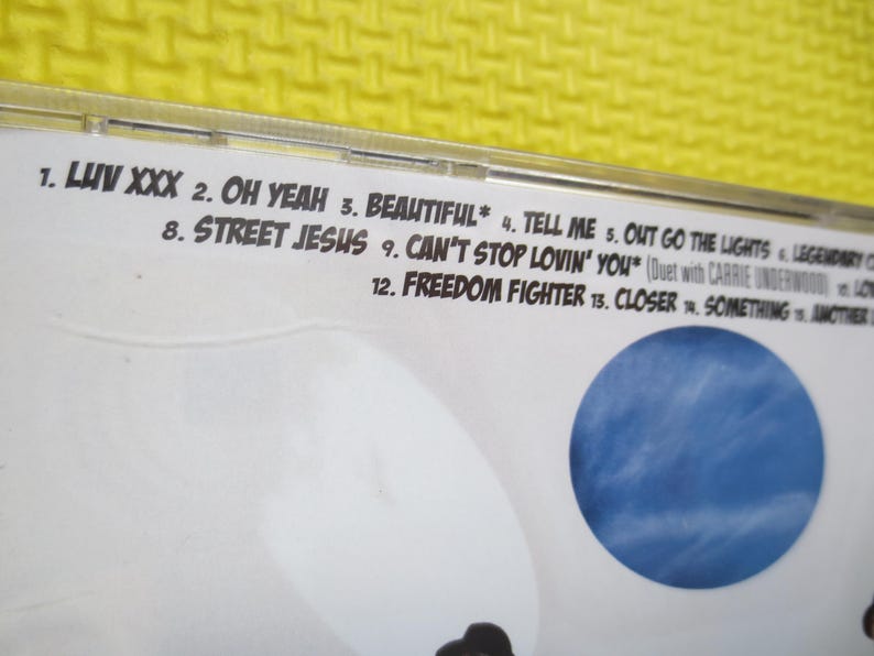 May include: A CD case with a white background and a blue sky image. The case lists the track titles for the album, including "Luv XXX", "Oh Yeah", "Beautiful", "Tell Me", "Out Go The Lights", "Street Jesus", "Can't Stop Lovin' You", "Freedom Fighter", "Closer", and "Something".