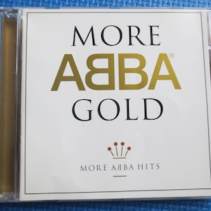Op de afbeelding: Een witte CD-doos met een gouden rand. De voorkant van de doos heeft de tekst "MORE ABBA GOLD" in zwarte en gouden letters. Onder de tekst staat een kleine rode kroon met vijf punten. De tekst "MORE ABBA HITS" is in zwart onder de kroon gedrukt.