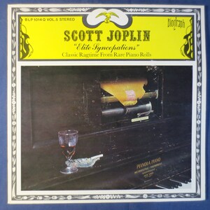 Peut inclure: Pochette d'album pour "Elite Syncopations" de Scott Joplin sur le label Biograph. La couverture présente un piano avec un verre de liquide et un cendrier. Le titre de l'album et le nom de l'artiste sont en texte gras sur fond jaune.