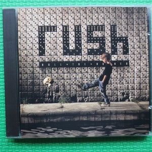 May include: A CD case with a black spine and a front cover featuring a child kicking a skull towards a wall with the word "DICE" written in large letters. The wall is made of small square tiles. The text "ROLL THE DICE" is written below the word "DICE".