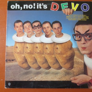 May include: A vinyl record cover for the band DEVO. The cover features a photo of five men dressed in white shirts and collars, with their heads replaced by potatoes. The text "oh, no! it's DEVO" is printed in large, colorful letters at the top of the cover.