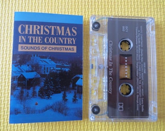 May include: A cassette tape with a blue and white cover featuring a snowy winter scene and the text "Christmas in the Country, Sounds of Christmas". The cassette tape is labeled "Christmas in the Country" and has a brown background.