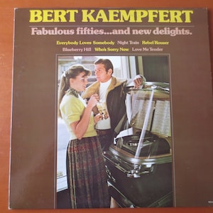 Può includere: Una copertina di un album in vinile vintage con una foto di una coppia in piedi davanti a un jukebox. L'album è intitolato "Bert Kaempfert: Fabulous fifties...and new delights." La copertina dell'album presenta i titoli delle canzoni "Everybody Loves Somebody", "Night Train", "Rebel Rouser", "Blueberry Hill", "Who's Sorry Now" e "Love Me Tender".