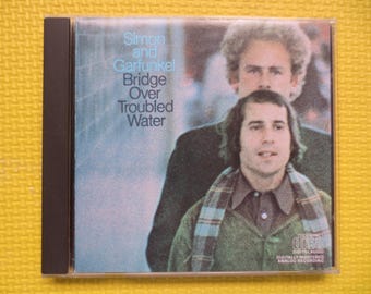 Può includere: Una copertina di un album vintage di Simon & Garfunkel. Il titolo dell'album è "Bridge Over Troubled Water". La copertina presenta una foto dei due musicisti, con Art Garfunkel che indossa una sciarpa.
