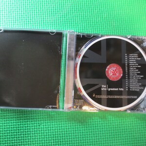 Puede incluir: Una caja de CD con un dise&ntilde;o de Union Jack en blanco y negro. El CD se titula "The Who | Greatest Hits".