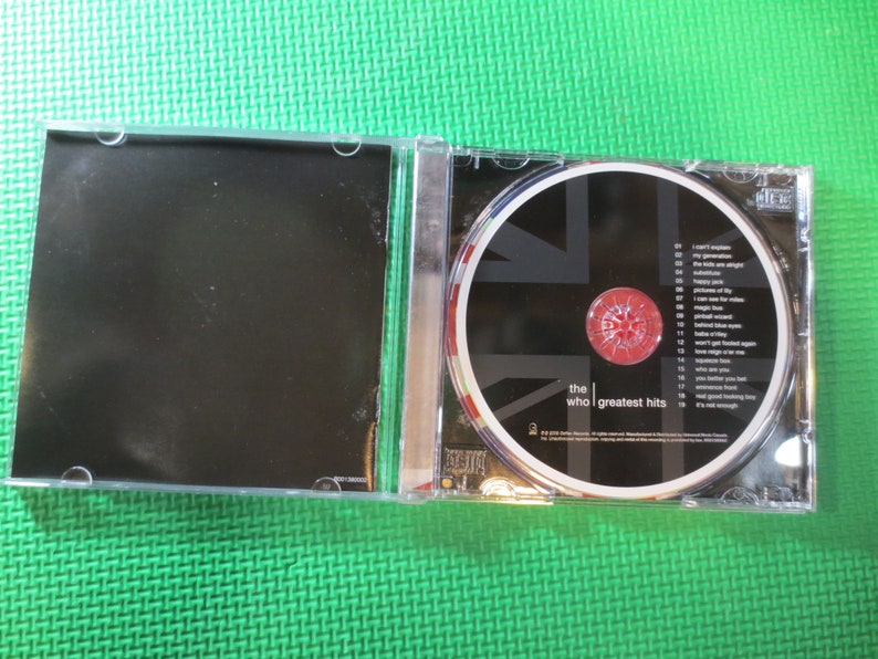 Puede incluir: Una caja de CD con un dise&ntilde;o de Union Jack en blanco y negro. El CD se titula "The Who | Greatest Hits".