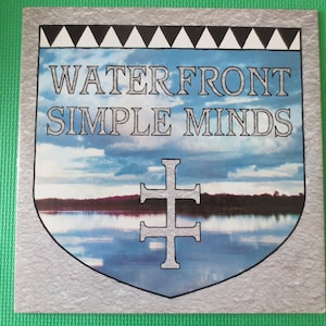 Peut inclure: Une pochette d'album de disque vinyle vintage représentant un paysage bleu et blanc avec une croix argentée au centre. Le titre de l'album est "WATERFRONT SIMPLE MINDS" en texte noir.
