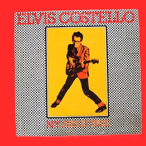 Peut inclure: Une pochette de disque vinyle carrée avec un panneau central jaune montrant une personne à lunettes jouant de la guitare électrique. La bordure est un motif en damier noir et blanc. Le texte ELVIS COSTELLO figure en haut et MY AIM IS TRUE en bas en rouge.