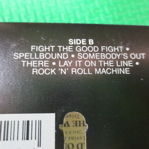 Puede incluir: Portada de &aacute;lbum en blanco y negro con el texto "SIDE B" y una lista de t&iacute;tulos de canciones: "Fight the Good Fight", "Spellbound", "Somebody's Out", "There", "Lay It on the Line" y "Rock 'n' Roll Machine".