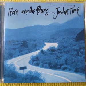 May include: A CD case with a blue and white image of a winding road through a desert landscape. The text "Here are the Blues... Justin Time" is written in black ink on the front of the case.