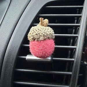 May include: A car air freshener shaped like an acorn with a textured, light brown natural cap and a round, fuzzy, bright pink felted body. The acorn is attached to a silver-colored clip that is secured onto the horizontal slats of a black car vent.