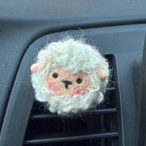 May include: A round, needle-felted sheep ornament with curly white wool, a peach-colored face, small black eyes, a dark nose, and pink blush on the cheeks. The sheep has two small, rounded ears and a textured, fluffy appearance throughout.