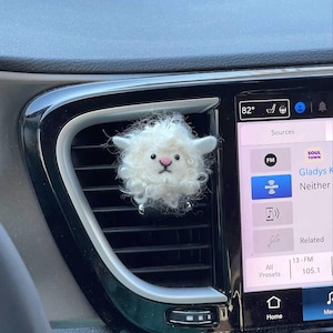 Könnte beinhalten: Ein weißer, flauschiger Schaf-Lufterfrischer für das Auto, der an einem Lüftungsgitter im Auto hängt. Das Schaf hat eine rosa Nase und schwarze Augen.