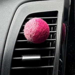 May include: A spherical, textured felt ball in a vibrant magenta pink color, attached to the horizontal slats of a black plastic automotive air conditioning vent. The felt material appears soft and fuzzy, contrasting with the smooth, dark surface.
