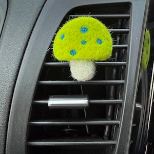 May include: A needle-felted mushroom ornament with a bright lime green cap featuring small blue polka dots and a white stem. The mushroom is attached to the horizontal slats of a black plastic car air vent, with a reflection visible in the side.