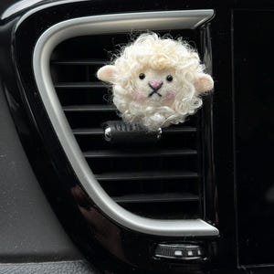 May include: A fluffy, white sheep car air vent clip. The sheep has a curly wool texture, pink cheeks, and a black embroidered cross for a mouth. The clip is attached to a black car vent.