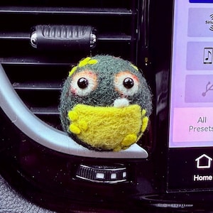 May include: A round, needle-felted creature ornament with a dark gray upper body and a bright yellow belly. It features two large, round black eyes with white and orange accents, a small white tooth, and yellow textured spots on its sides.