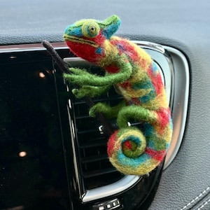 May include: A colorful felt chameleon perched on a car vent. The chameleon features a vibrant mix of red, green, blue, and yellow. It is holding a small, dark-colored branch, adding a touch of realism to the handcrafted design.