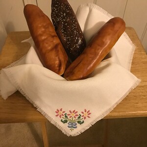 Può includere: Tre pagnotte di pane, una scura e due chiare, sono esposte in un cesto di stoffa bianca con un ricamo floreale. Il cesto è su un tavolo di legno.