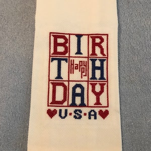 Pode incluir: Toalha de m&atilde;o creme com um design bordado em ponto cruz. O design apresenta as palavras "BIRTHDAY" e "USA" em vermelho e azul, com um pequeno "Happy" no centro. Dois cora&ccedil;&otilde;es vermelhos ladeiam a palavra "USA".