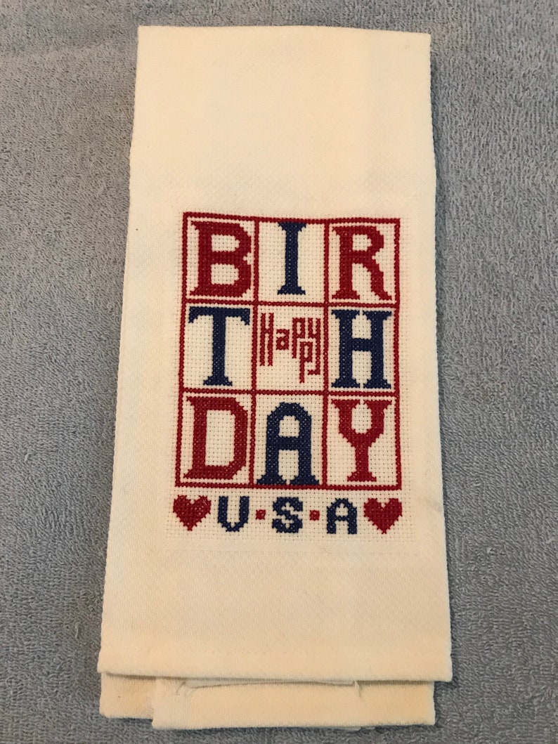 Pode incluir: Toalha de m&atilde;o creme com um design bordado em ponto cruz. O design apresenta as palavras "BIRTHDAY" e "USA" em vermelho e azul, com um pequeno "Happy" no centro. Dois cora&ccedil;&otilde;es vermelhos ladeiam a palavra "USA".