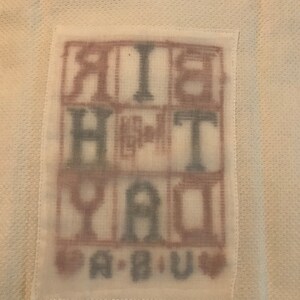 Pode incluir: Um tecido branco com um patch retangular com as palavras "BIRTHDAY" e "A B U" em um estilo de letras de bloco. As letras est&atilde;o em uma mistura de marrom e azul, com s&iacute;mbolos de cora&ccedil;&atilde;o.