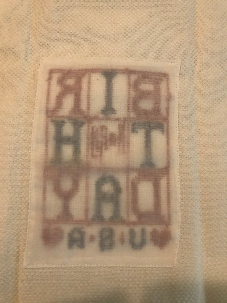 Pode incluir: Um tecido branco com um patch retangular com as palavras "BIRTHDAY" e "A B U" em um estilo de letras de bloco. As letras est&atilde;o em uma mistura de marrom e azul, com s&iacute;mbolos de cora&ccedil;&atilde;o.