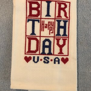Pode incluir: Uma toalha dobrada, de cor creme, bordada em ponto cruz com as palavras "BIRTHDAY" e "USA" em vermelho e azul. A palavra "Happy" est&aacute; no centro. Dois cora&ccedil;&otilde;es vermelhos ladeiam a palavra "USA".