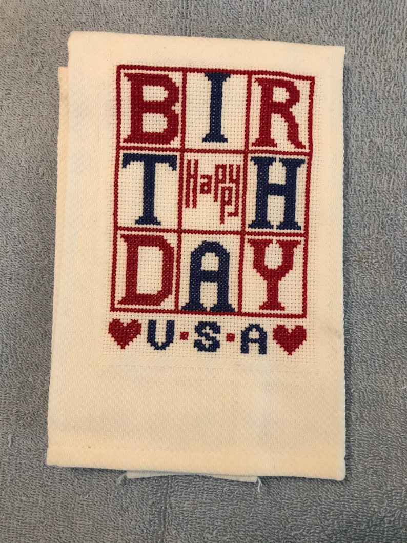 Pode incluir: Uma toalha dobrada, de cor creme, bordada em ponto cruz com as palavras "BIRTHDAY" e "USA" em vermelho e azul. A palavra "Happy" est&aacute; no centro. Dois cora&ccedil;&otilde;es vermelhos ladeiam a palavra "USA".