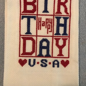 Pode incluir: Uma toalha branca bordada em ponto cruz com as palavras "BIRTHDAY" em vermelho e azul, e "Happy" em vermelho. Abaixo, diz "U.S.A" com cora&ccedil;&otilde;es vermelhos. A toalha est&aacute; dobrada.