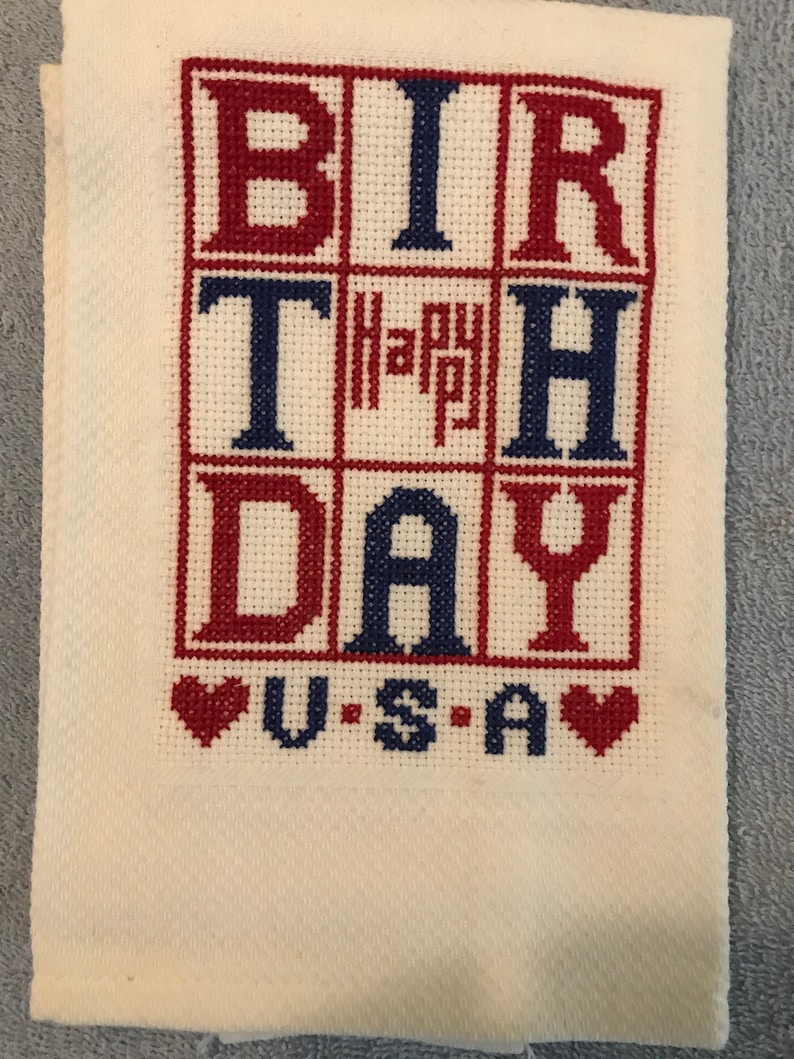Pode incluir: Uma toalha branca bordada em ponto cruz com as palavras "BIRTHDAY" em vermelho e azul, e "Happy" em vermelho. Abaixo, diz "U.S.A" com cora&ccedil;&otilde;es vermelhos. A toalha est&aacute; dobrada.