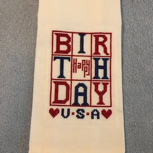 Pode incluir: Toalha de m&atilde;o creme com texto bordado em ponto cruz "BIRTHDAY USA" em vermelho e azul. A palavra "Happy" est&aacute; no centro. Dois cora&ccedil;&otilde;es vermelhos em ambos os lados de "USA".