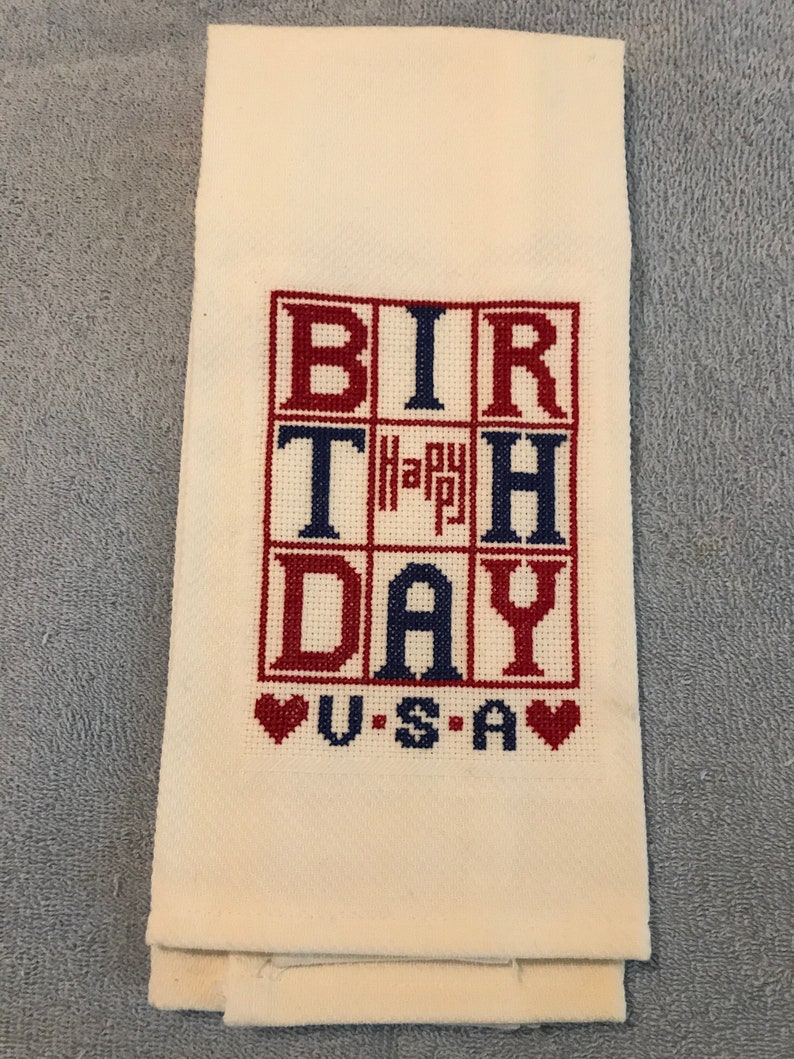 Pode incluir: Toalha de m&atilde;o creme com texto bordado em ponto cruz "BIRTHDAY USA" em vermelho e azul. A palavra "Happy" est&aacute; no centro. Dois cora&ccedil;&otilde;es vermelhos em ambos os lados de "USA".