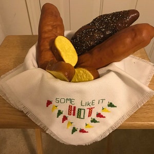 May include: A white cloth napkin with a fringed edge and embroidered text "Some Like It Hot" with red, green, and yellow peppers. The napkin is in a white ceramic bowl filled with bread rolls, including a pretzel roll and a long roll with black sesame seeds.