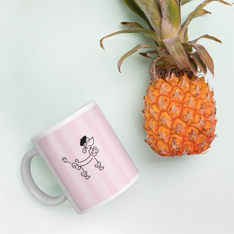 May include: White ceramic mug with a pink and white striped design. A black and white illustration of a poodle wearing a beret is on the mug. A pineapple is in the background.