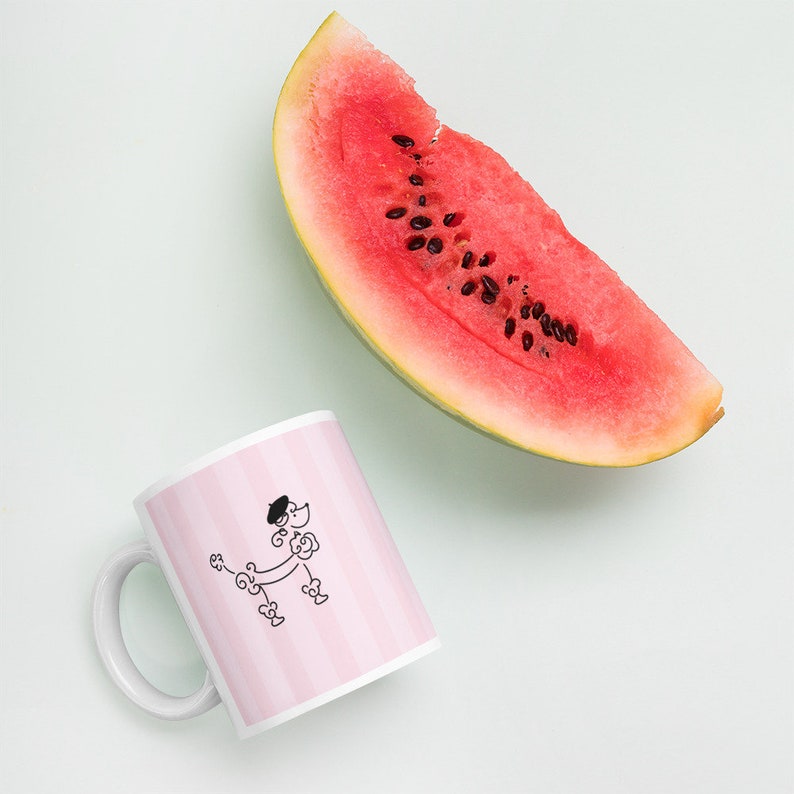 May include: A white ceramic mug with a pink and white striped design. A black and white illustration of a poodle wearing a beret is on the mug. A slice of watermelon is in the background.