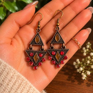 May include: A pair of bronze-toned earrings with a geometric design. Each earring features a triangle shape with a central amber-colored stone and red jewel accents. The earrings are held in a hand, showcasing their intricate details.
