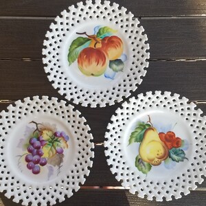 May include: Three white porcelain dessert plates with scalloped edges and a delicate lattice design. Each plate features a hand-painted fruit motif: apples, grapes, and a pear.