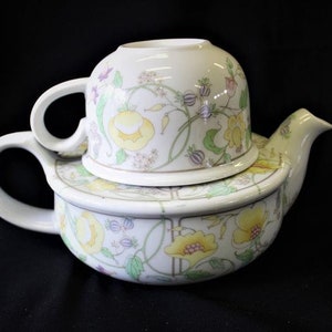 May include: A white teapot with a floral design in shades of yellow, green, and pink. The teapot has a removable lid that doubles as a cup.