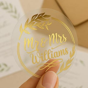 Peut inclure: Un autocollant rond transparent avec des lettres dorées et un motif floral doré. Le texte indique "Mr & Mrs Williams". L'autocollant est tenu dans une main, avec de la papeterie de mariage en arrière-plan.