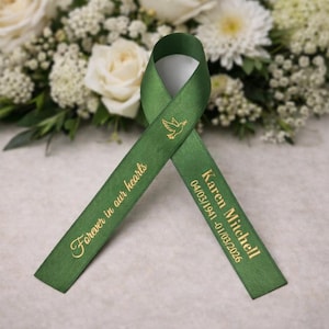 May include: A green satin ribbon with gold text and a dove graphic. The text reads "Forever in our hearts" and "Karen Mitchell 04/03/1941-01/03/2026". The ribbon is displayed in front of a floral arrangement of white roses and chrysanthemums.