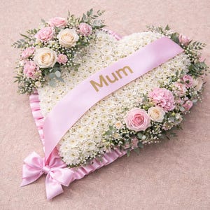 May include: A heart-shaped floral tribute with white and pink flowers, a pink ribbon with the word "Mum", and a pink bow. The arrangement includes roses, carnations, and greenery.