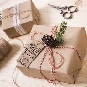 May include: Two brown paper wrapped gifts tied with twine. One gift has a white lace ribbon and the other has a red and white twine with a pine cone and evergreen sprig. Both gifts have a tag that says "North Pole Post Special Delivery" with a reindeer and sleigh graphic.