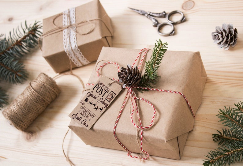 May include: Two brown paper wrapped gifts tied with twine. One gift has a white lace ribbon and the other has a red and white twine with a pine cone and evergreen sprig. Both gifts have a tag that says "North Pole Post Special Delivery" with a reindeer and sleigh graphic.