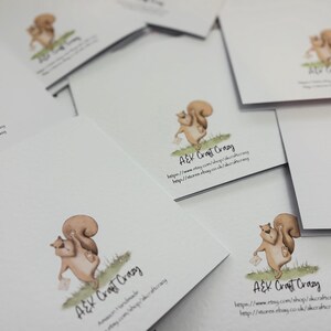 May include: A collection of white greeting cards featuring a watercolor illustration of a squirrel holding a letter. The squirrel is depicted in shades of brown and tan, standing on green grass. The cards have the text "A&K Craft Crazy" and website information.