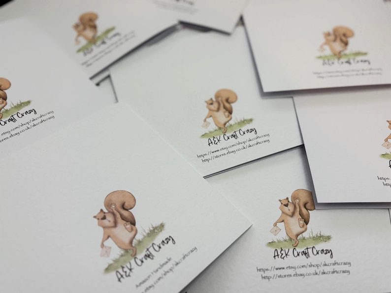 May include: A collection of white greeting cards featuring a watercolor illustration of a squirrel holding a letter. The squirrel is depicted in shades of brown and tan, standing on green grass. The cards have the text "A&K Craft Crazy" and website information.