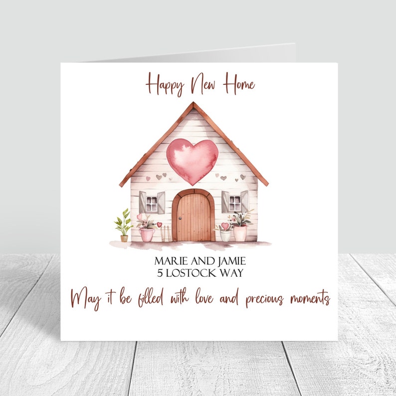 May include: A white house with a pink heart on the front, surrounded by flowers and plants. The text "Happy New Home" is at the top of the card, and the text "Marie and Jamie 5 Lostock Way May it be filled with love and precious moments" is at the bottom.