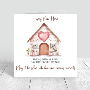 May include: A watercolor illustration of a white house with a pink heart on the front. The house has a brown door and windows. There are plants in pots on either side of the house. The text "Happy New Home" is at the top of the card. The text "SIMON, EMMA & LEAH 34 GREEN BELLE AVENUE" is below the house. The text "May it be filled with love and precious moments" is at the bottom of the card.