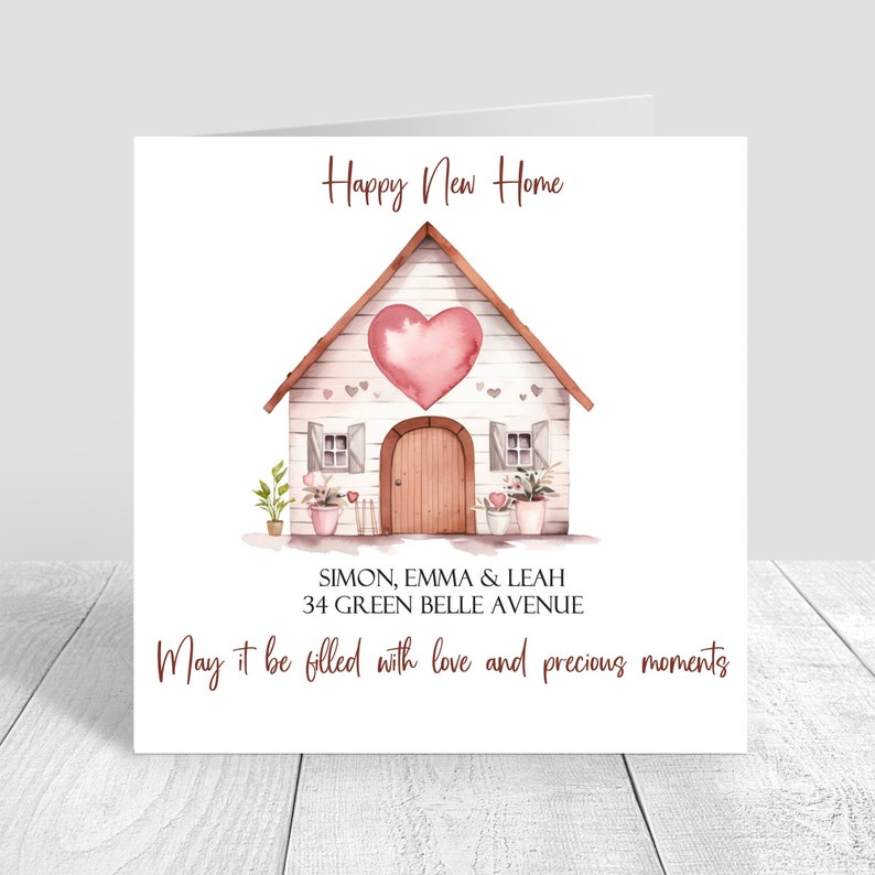 May include: A watercolor illustration of a white house with a pink heart on the front. The house has a brown door and windows. There are plants in pots on either side of the house. The text "Happy New Home" is at the top of the card. The text "SIMON, EMMA & LEAH 34 GREEN BELLE AVENUE" is below the house. The text "May it be filled with love and precious moments" is at the bottom of the card.